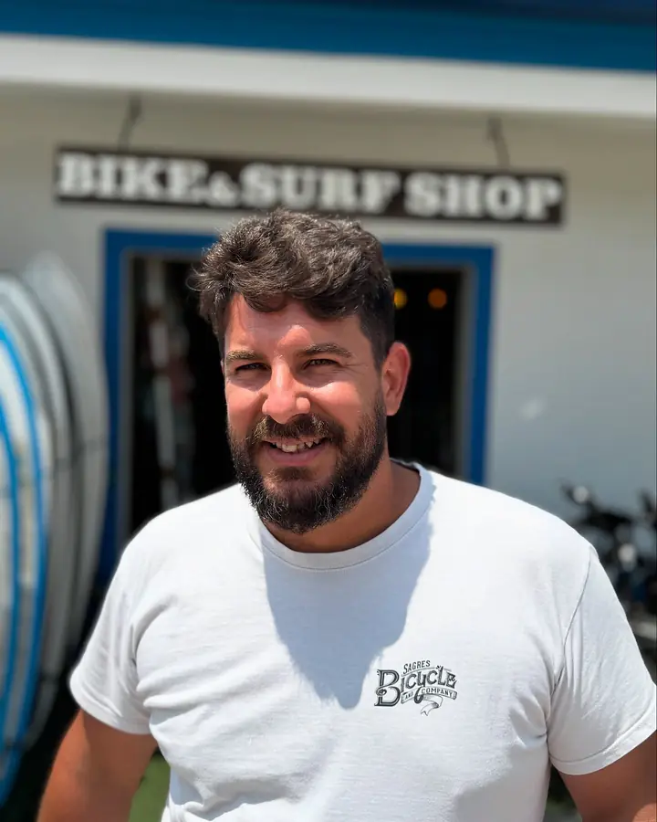 Bruno Silva, dono da Sagres Bicycle and Company, a sorrir à porta da loja, de t-shirt com o logótipo, por baixo da placa Bike & Surf Shop.
