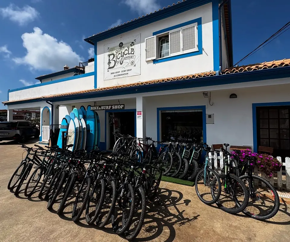 The Sagres Bicycle and Company shop on the EN 268: a white building with blue trim, the logo sign above the door, surfboards stacked by the entrance and a row of rental bikes outside.