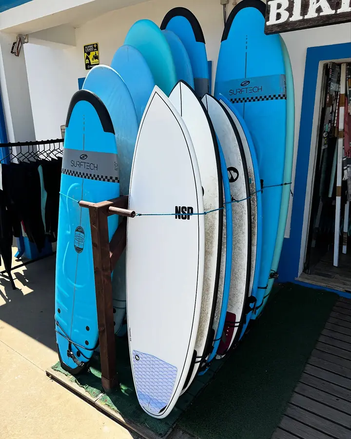 Rental surfboards in the rack outside the shop: blue Surftech soft-tops and white NSP shortboards, with wetsuits hanging alongside.